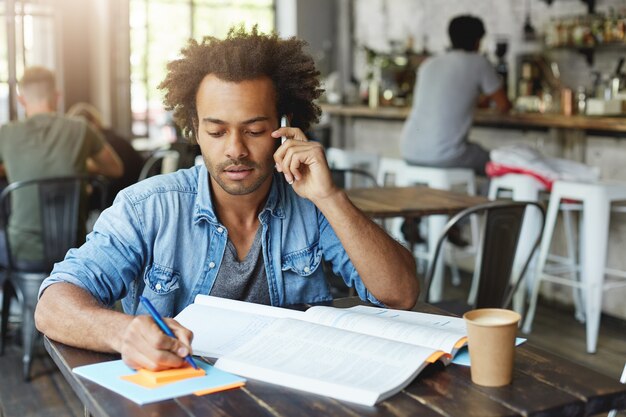 Retrato interior de atractivo estudiante de posgrado universitario afroamericano de moda hablando por teléfono inteligente con su supervisor de investigación mientras trabaja en papel de diploma, sentado en la mesa de café