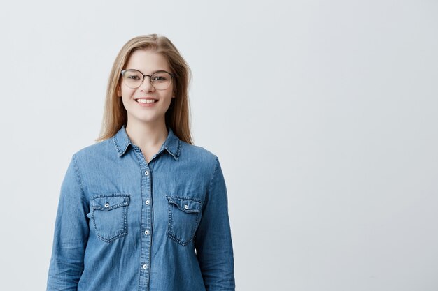 Retrato interior de una bella joven rubia europea con el pelo liso, con gafas elegantes, sonriendo, mostrando sus dientes blancos a la cámara, sintiéndose feliz y despreocupada en su primer día