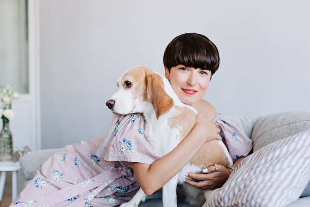 Retrato de interior de chica atractiva con peinado corto abrazando cachorro grande con ojos negros