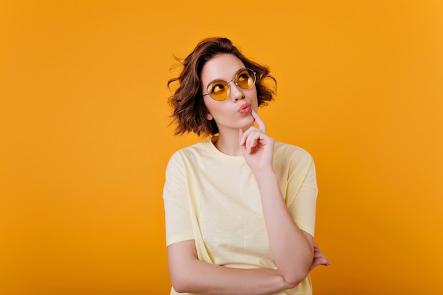 Retrato de interior de una chica morena pensativa en camiseta de color amarillo claro. Alegre mujer de pelo corto con gafas de sol mirando hacia arriba y pensando en algo.