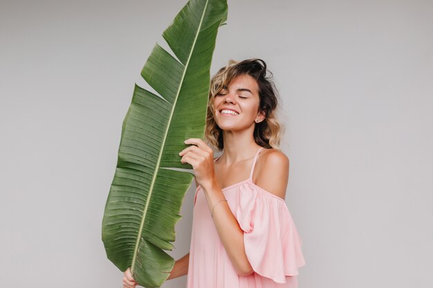 Retrato interior de una chica refinada posando con los ojos cerrados cerca de una planta exótica. adorable mujer de pelo corto sosteniendo una gran hoja verde y sonriendo.