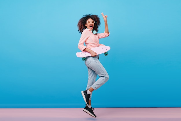 Retrato interior de cuerpo entero de una niña africana segura en camisa rosa con patineta. Entusiasta mujer negra con peinado rizado posando con interior azul.