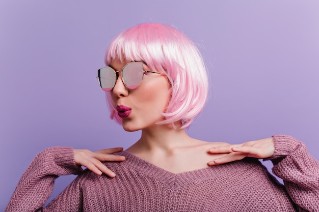 Retrato de interior de dama encantadora con gafas brillantes y suéter de punto de pie en la pared púrpura. Foto de chica glamorosa con peluca rosa posando con expresión de cara de besos.