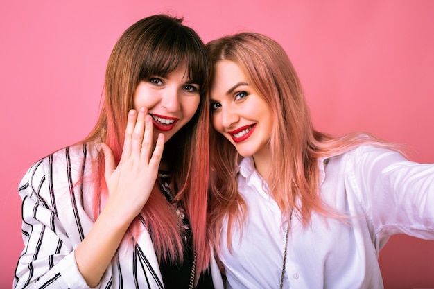 Retrato interior de dos felices mejores amigas hermanas mujeres, vistiendo ropa de moda en blanco y negro y pelos rosados, haciendo selfie, disfrutando del tiempo juntos