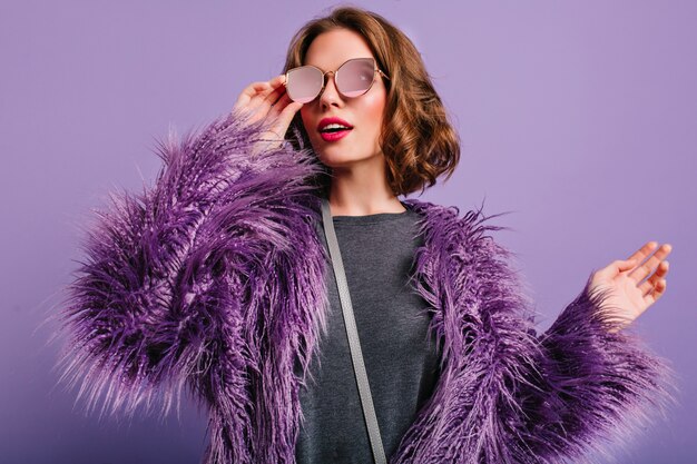 Retrato de interior de elegante modelo femenino europeo en gafas posando sobre fondo púrpura