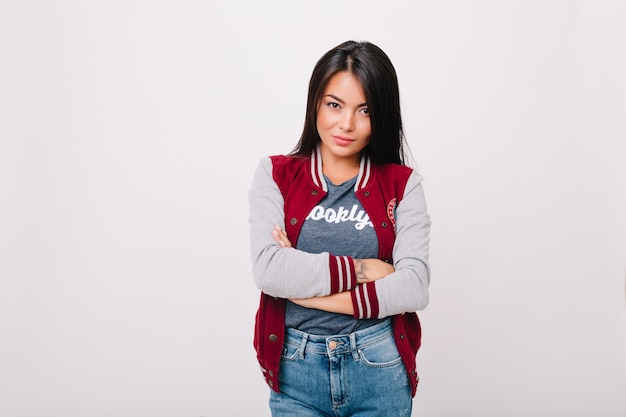 Retrato interior de una encantadora chica asiática. Linda estudiante de pelo oscuro en bombardero posando con los brazos cruzados aislados.