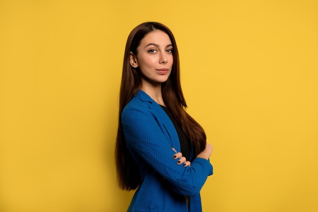 Retrato de interior de exitosa mujer joven con cabello largo oscuro vistiendo chaqueta azul posando con los brazos cruzados sobre la pared amarilla