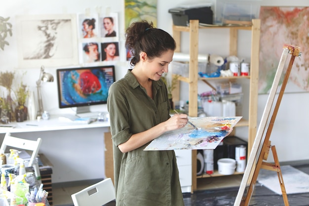 Retrato interior de feliz joven artista emocionada en camisa de color militar con paleta y pincel mientras trabajaba en la pintura en su taller, de pie delante del caballete y sonriendo