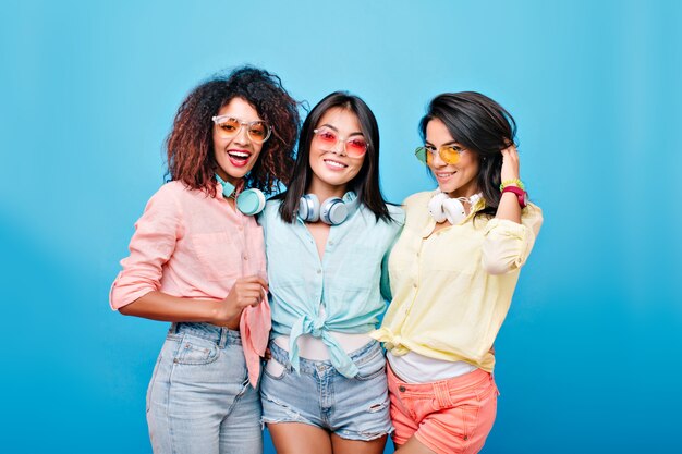 Retrato interior de grupo de fascinantes amigos internacionales con atuendos coloridos y gafas de sol brillantes. Señoras morenas sonrientes de diferentes etnias posando juntas.