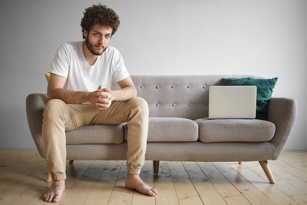 Retrato interior de un hombre barbudo joven autónomo de moda en ropa casual que trabaja desde casa, usando wifi en un dispositivo electrónico portátil, sentado en el sofá en la sala de estar con una computadora portátil genérica