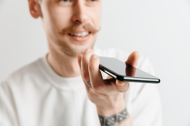 Retrato interior de un joven atractivo aislado en un espacio gris, sosteniendo un teléfono inteligente, usando el control de voz, sintiéndose feliz y sorprendido