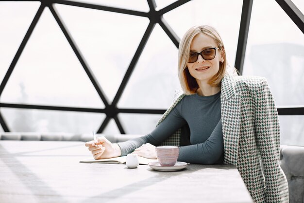 Retrato interior de una joven empresaria sentada en un café y escribiendo documentos. Chica rubia con chaqueta y gafas de sol