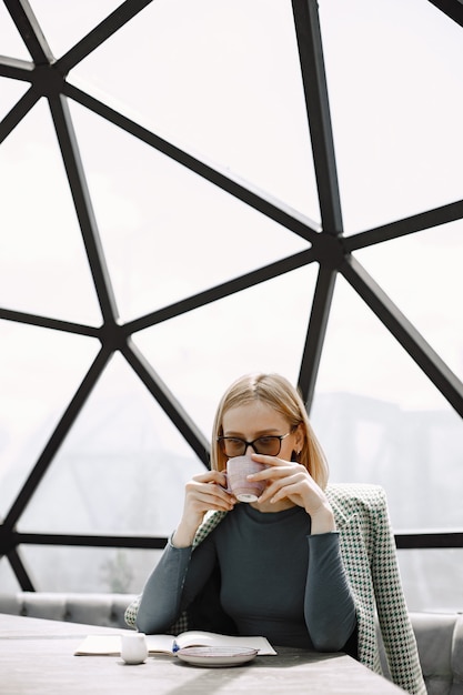 Retrato interior de una joven empresaria sentada en un café y escribiendo documentos. Chica rubia con chaqueta y gafas de sol