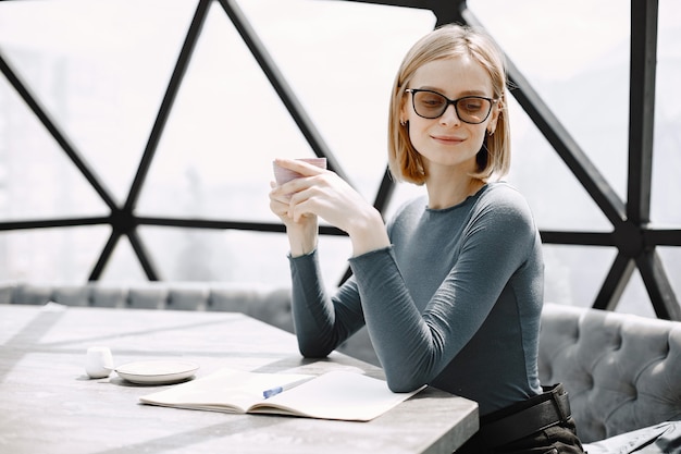 Retrato interior de una joven empresaria sentada en un café y tomando un café. Chica rubia con chaqueta y gafas de sol