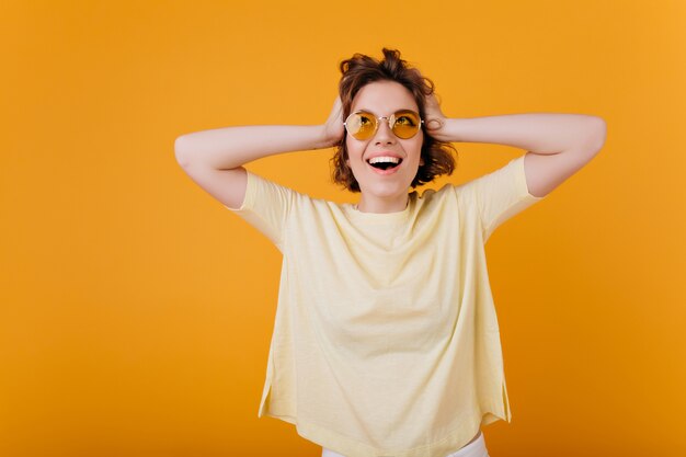 Retrato interior de una jovencita emocionada en una camiseta amarilla de gran tamaño tocando su cabeza. emocional niña caucásica feliz riendo.