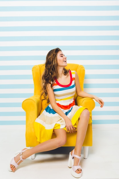Retrato de interior de una linda chica inspirada con sandalias de tacón alto y vestido de rayas de colores. Agraciada joven con piel bronceada en sillón amarillo de pie en su habitación y riendo.