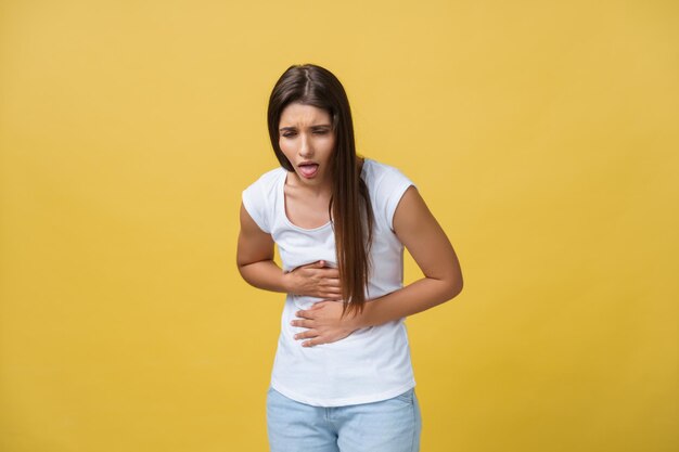 Retrato interior de una linda chica parada con las manos cruzadas en el vientre sintiéndose incómoda o sufriendo de dolor mientras mira a un lado de pie contra un fondo amarillo La mujer tiene dolor de estómago