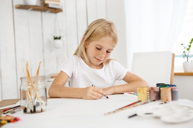 Retrato interior de linda niña rubia con pecas dibujando con lápices de colores en la hoja de papel