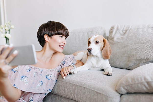 Retrato de interior de maravillosa chica morena haciendo selfie con perro beagle acostado en el sofá