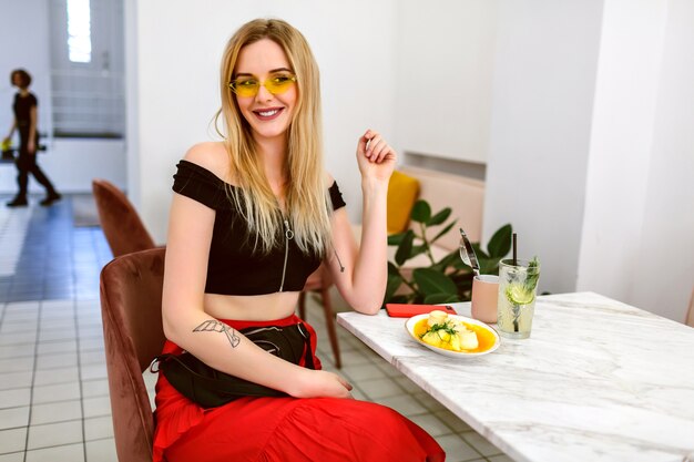 Retrato interior de moda de mujer rubia hipster de moda joven posando en el café moderno hipster, hora del desayuno por la mañana.