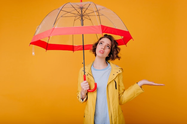 Retrato interior del modelo femenino joven trastornado en impermeable del otoño. Foto de dama rizada triste de pie bajo el paraguas de moda y mirando hacia arriba.