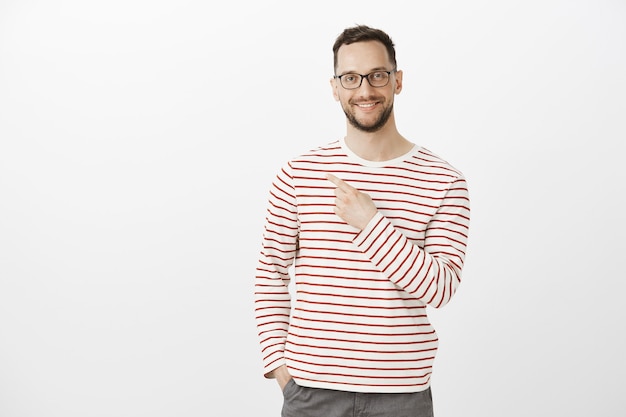 Retrato interior de un modelo de hombre guapo y complacido con gafas, sonriendo ampliamente y apuntando hacia atrás o hacia la esquina superior izquierda