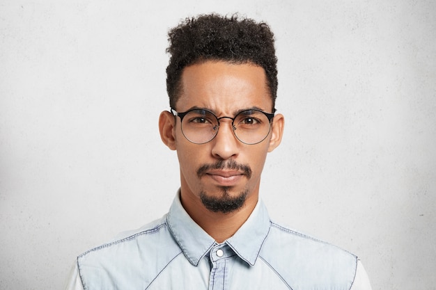 Retrato interior de modelo masculino molesto descontento disgustado con peinado de moda, bigote, barba, usa gafas redondas