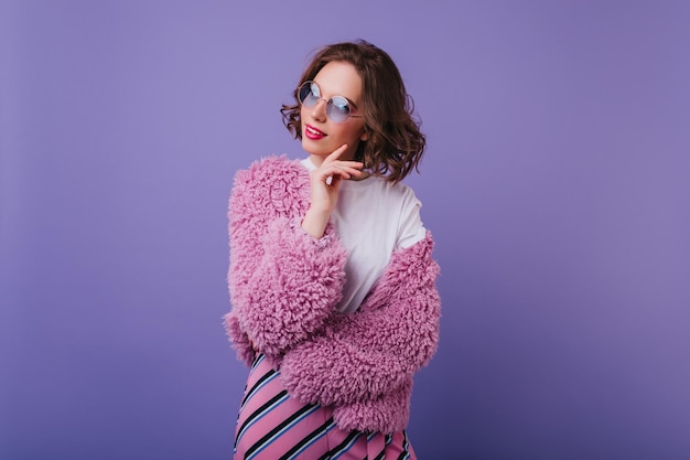 Retrato interior de una mujer caucásica elegante que viste una chaqueta brillante de moda Chica soñadora con gafas de sol posando con placer en un abrigo de piel sobre fondo morado