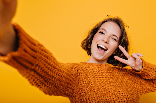 Retrato interior de mujer dichosa en ropa tejida haciendo selfie y riendo sobre fondo brillante