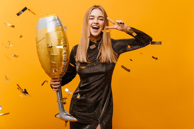 Retrato interior de mujer encantadora riendo con copa de vino posando en amarillo. Hermosa mujer de cumpleaños en vestido de pie bajo confeti y riendo.