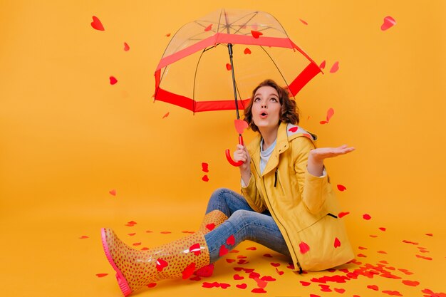 Retrato interior de una mujer hermosa conmocionada mirando la lluvia del corazón. Disparo de estudio de niña alegre con paraguas aislado en pared brillante en el día de San Valentín.