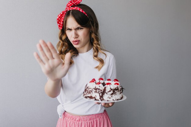 Retrato de interior de mujer infeliz lleva cinta roja posando con pastel. cumpleañera sosteniendo pastel.