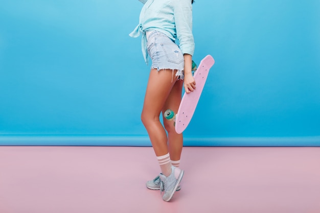 Retrato interior de mujer joven deportiva con piel bronceada disfrutando junto a la pared azul brillante. Chica bien formada con longboard usa zapatos de moda y calcetines lindos.