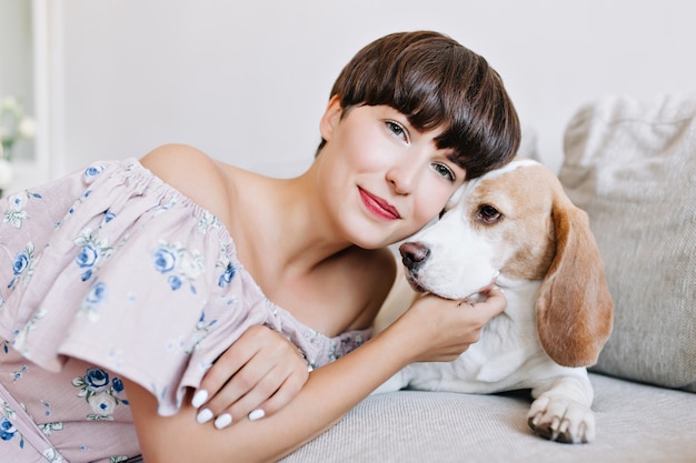 Retrato interior de mujer joven romántica con peinado corto de moda posando tocando cachorro beagle y sonriendo