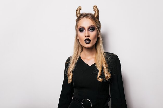 Retrato de interior de mujer rizada de pelo largo posando después del carnaval. Chica vampiro interesada de pie en la pared blanca.