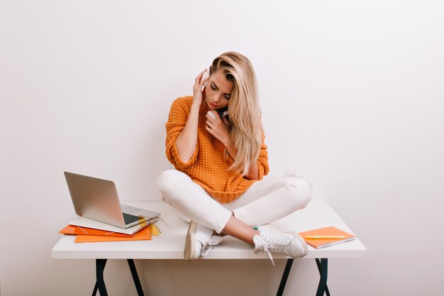 Retrato interior de mujer rubia cansada sentada en la mesa con las piernas cruzadas y sosteniendo los auriculares