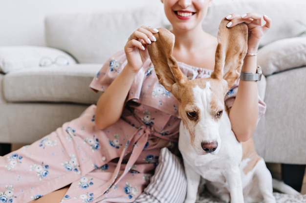 Retrato interior de una mujer sonriente en un romántico vestido rosa con un lindo cachorro beagle en primer plano. Increíble chica con manicura blanca jugando con orejas de perro y riendo