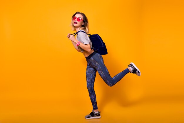 Retrato de interior de niña riendo jugando con músculos sobre fondo amarillo. Mujer joven bien formada en leggings y camiseta gris sosteniendo una botella de agua y haciendo deporte.