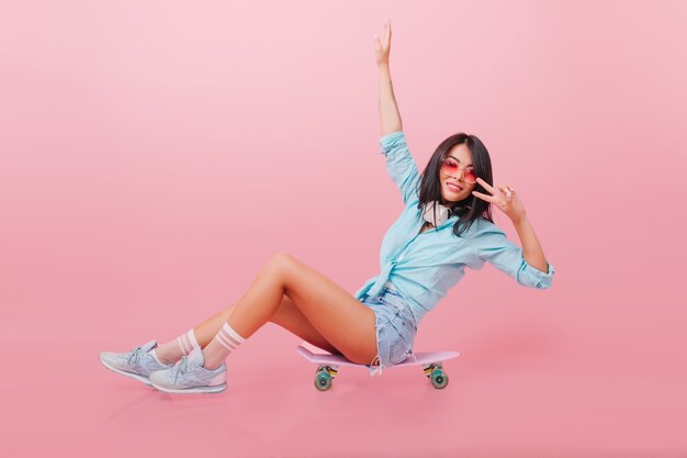 Retrato interior de una relajada chica morena sentada en longboard y agitando las manos. Modelo femenino asiático entusiasta en zapatillas lindas posando con patineta con interior rosa.