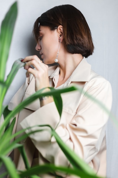 Retrato interior suave de mujer caucásica suave con traje beige sin sujetador, posando detrás de una planta tropical de palma, gris.