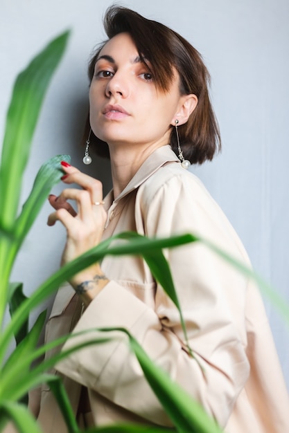 Retrato interior suave de mujer caucásica suave con traje beige sin sujetador, posando detrás de una planta tropical de palma, gris.