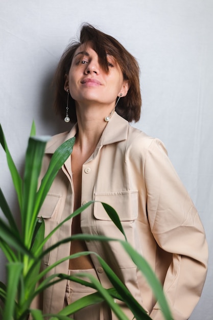 Retrato interior suave de mujer caucásica suave con traje beige sin sujetador, posando detrás de una planta tropical de palma, gris.