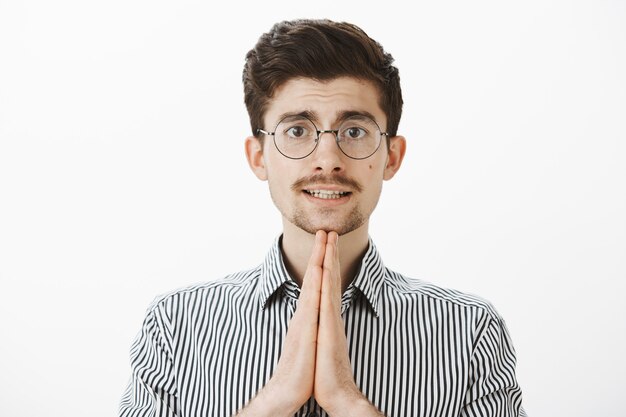 Retrato interior de un tímido chico europeo común con gafas de moda, tomados de la mano en oración y pidiendo un favor o disculpándose, esperando perdón con una expresión linda, de pie sobre una pared gris