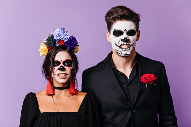 Retrato interior de zombies enojados aislados sobre fondo púrpura. Divertida pareja emocional en disfraces de muertos posando con expresión de miedo.