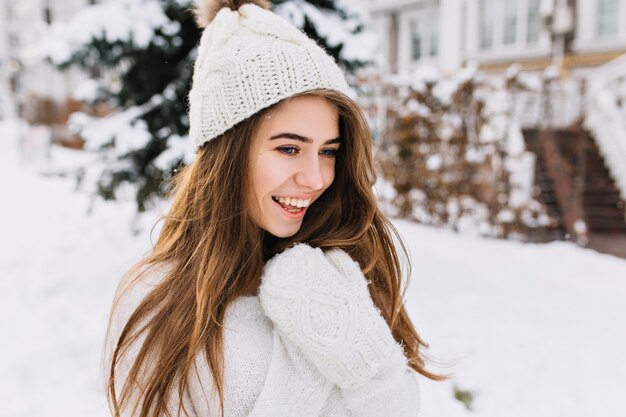 Retrato de invierno acogedor de mujer joven alegre de moda con cabello largo morena caminando en la calle llena de nieve. Emociones verdaderas positivas sorprendidas, cálidos guantes de lana blanca, gorro de punto.