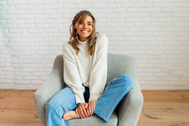 Retrato de invierno en el interior de una mujer bonita en un acogedor suéter sentado en un sofá moderno en casa.