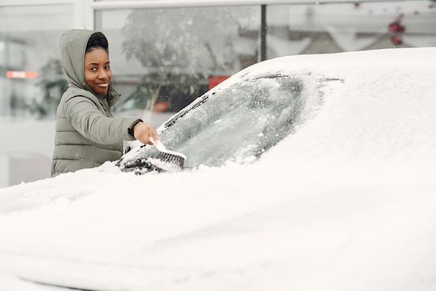 Retrato de invierno de mujer africana, limpiar la nieve de un coche. Mujer con chaqueta verde.