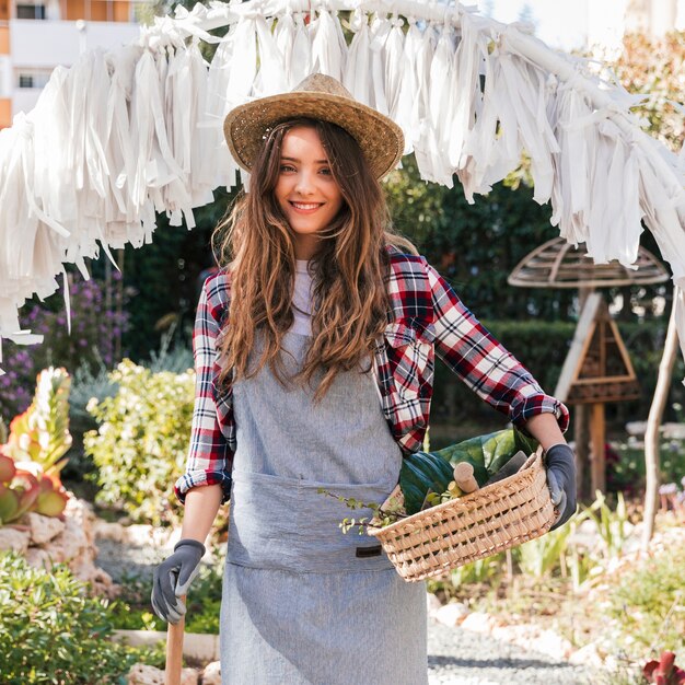 Retrato de un jardinero de sexo femenino joven sonriente que sostiene la herramienta y la cesta que cultivan un huerto