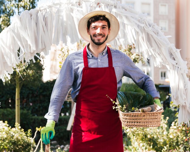 Retrato de un jardinero de sexo masculino joven sonriente que sostiene la herramienta y la cesta que cultivan un huerto que miran la cámara