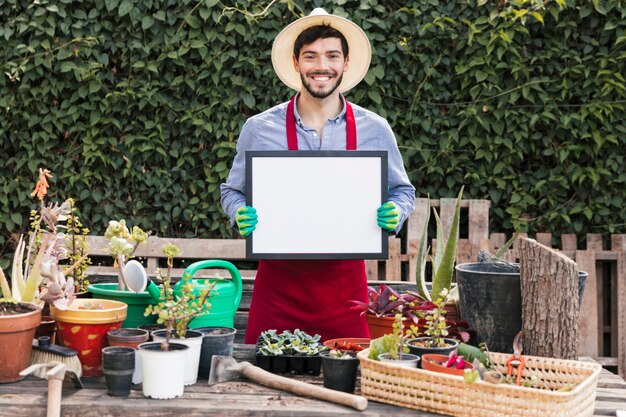 Retrato de un jardinero de sexo masculino sonriente que lleva a cabo el marco blanco delante de las plantas en conserva en la tabla